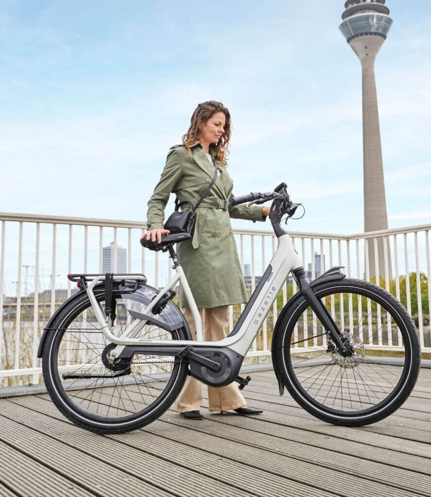 A woman in a green trench coat and work clothes stands beside a white Gazelle e-bike on a wooden deck. She holds the handlebars and seat, looking downward with a thoughtful calm expression.