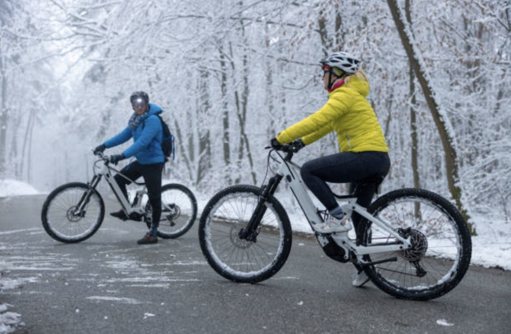 Two cyclists in winter jackets ride electric bikes on a snow-covered path in a forest. Snow covers the trees, creating a beautiful winter scene as they appear to be enjoying a peaceful outdoor ride with their appropriate winter gear