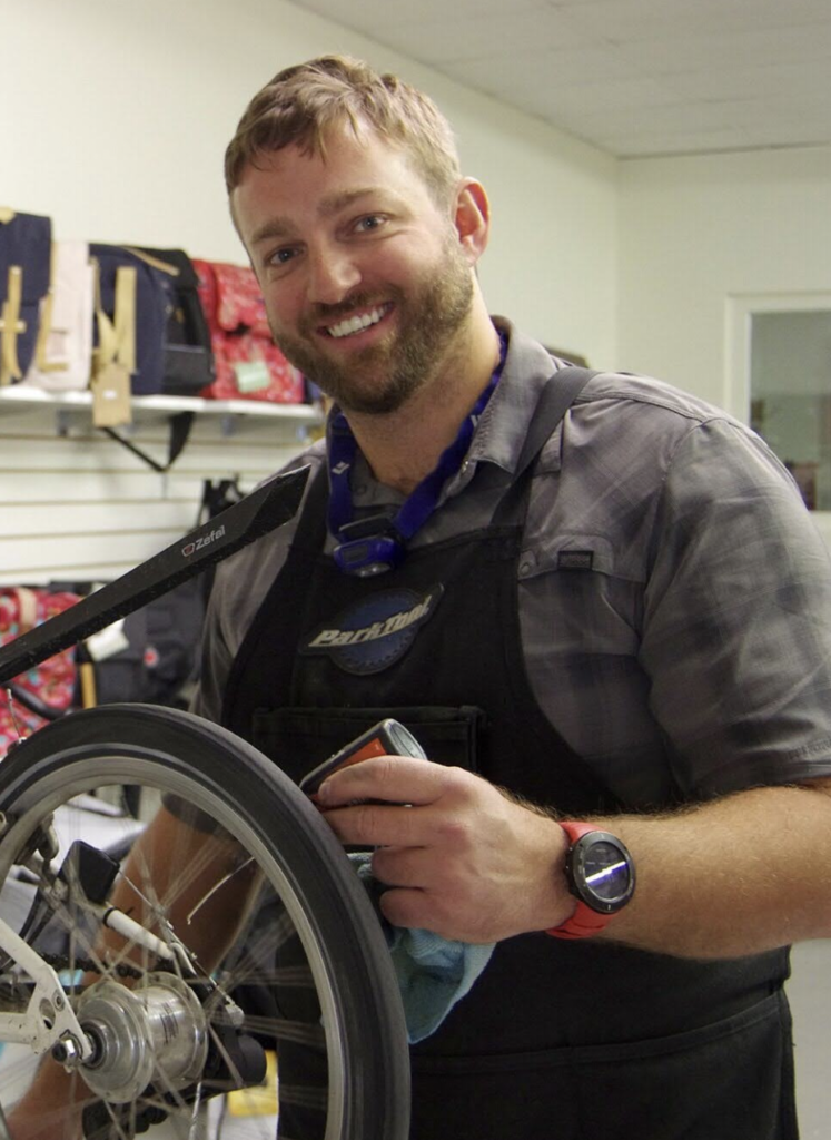 A smiling mechanic, holding a cloth as he works on a bicycle wheel. He wears a gray shirt, a black apron, and a blue lanyard around his neck, with various tools visible in the background.