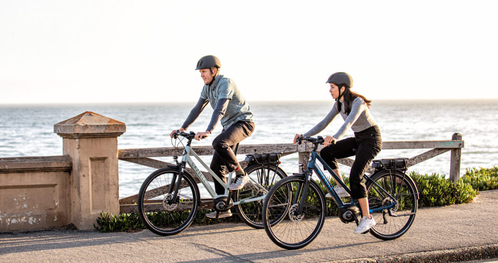 A woman and man riding their gazelle e-bikes on a bridge beside a big body of water on a nice sunny